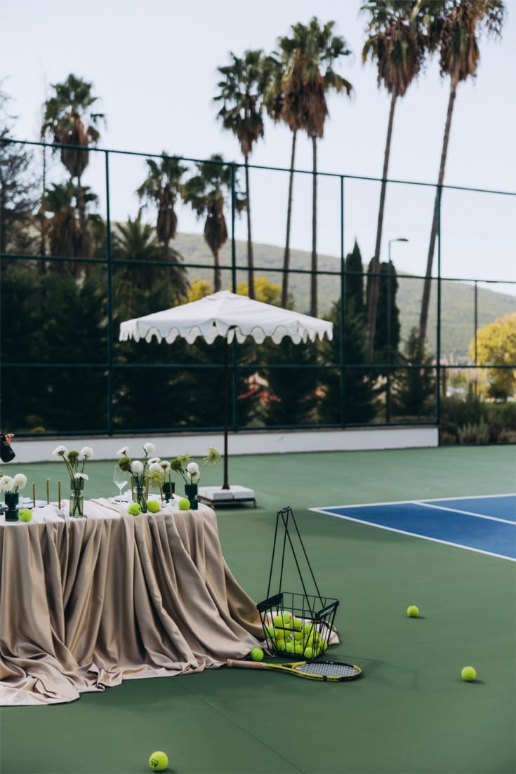A courtside table under a scalloped umbrella, balls and a racket on the court